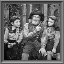 George "Gabby" Hayes in his usual battered and ragged, down to the very obvious large "patch" on the right knee, not-quite-a-cowboy outfit is explaining something to two children - boy on Gabby's right, girl on his left - both dressed in "Western-style" outfits in keeping with the theme of Gabby's television show.